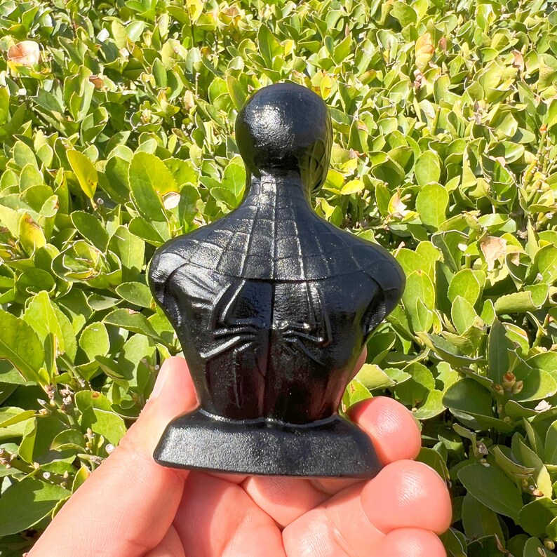 May include: A black bust of Spider-Man, viewed from the back, with the iconic spider emblem visible. The sculpture is set against a backdrop of green foliage, highlighting the detailed texture of the superhero's suit.