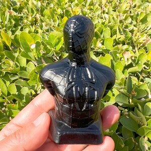 May include: A black bust of Spider-Man, featuring the iconic spider emblem on the chest and a detailed web pattern. The sculpture is glossy and stands on a rectangular base, showcasing the superhero's likeness.