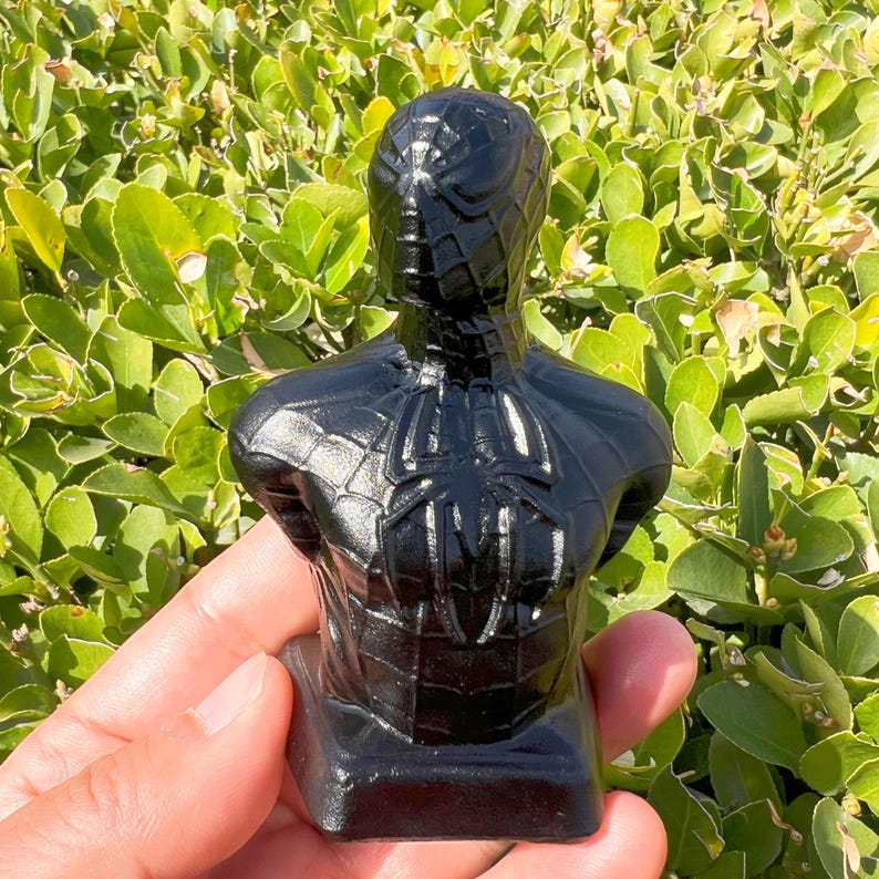 May include: A black bust of Spider-Man, featuring the iconic spider emblem on the chest and a detailed web pattern. The sculpture is glossy and stands on a rectangular base, showcasing the superhero's likeness.