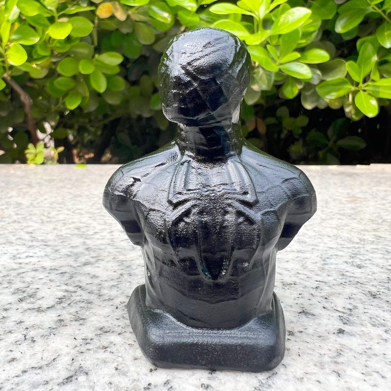 May include: A black bust of a superhero with a spider emblem on the back. The sculpture is made of a dark, glossy material and is set against a backdrop of green foliage.