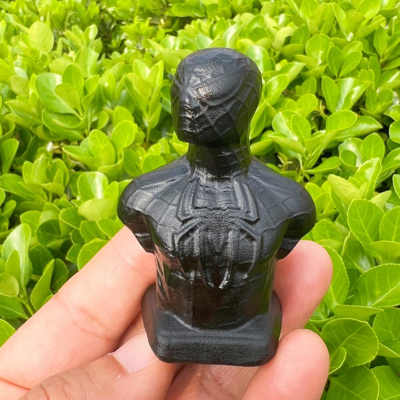 May include: A black bust of a superhero with a spider emblem on the chest. The sculpture is held in a hand against a backdrop of green foliage. The bust is a detailed representation of the character.