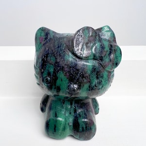 May include: A polished stone figurine of a cartoon cat character, featuring a bow and rounded features. The sculpture is primarily green with dark blue and purple veining, giving it a unique, natural appearance. The figurine is set against a white background.
