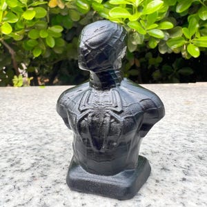 May include: A black bust of a superhero with a spider emblem on the chest. The sculpture is made of a dark material and is set against a background of green foliage.