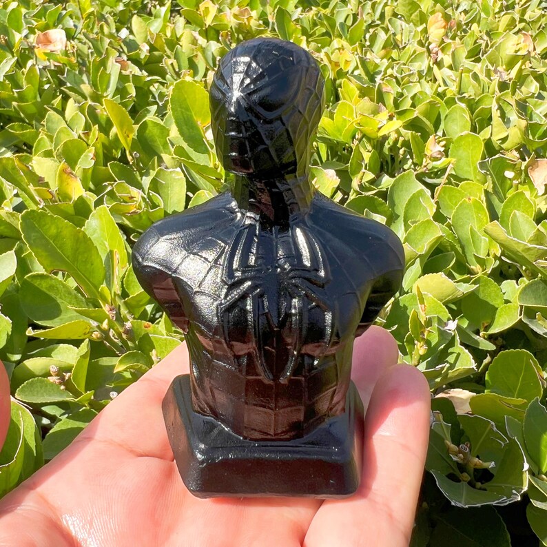 May include: A black bust of Spider-Man, featuring the character's iconic spider-web design and spider emblem on the chest. The sculpture is held in a hand, with a backdrop of green foliage.