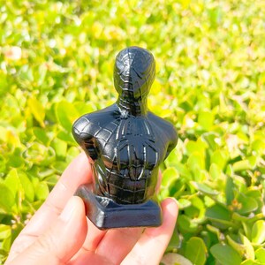 May include: A black bust of Spider-Man, featuring a detailed design of the superhero's suit. The sculpture is set against a backdrop of green foliage, highlighting the glossy finish of the figure. The bust is mounted on a rectangular base.