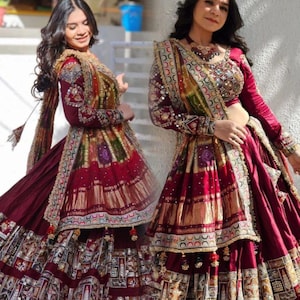 Puede incluir: Un conjunto de lehenga choli color granate que incluye una blusa de manga larga con peplum y una falda voluminosa. La prenda está ricamente adornada con intrincados parches multicolores, bordados dorados metálicos y pequeños espejos.