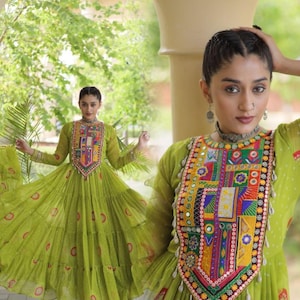May include: A lime green long-sleeved dress featuring a tiered skirt with a subtle red and white pattern. The bodice is adorned with a large, rectangular panel of multi-colored embroidery, small circular mirrors, and white shell accents.