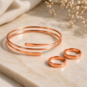 Pode incluir: Uma pulseira tipo bracelete em tom de ouro rosa com acabamento liso e polido, acompanhada por dois anéis a condizer em tom de ouro rosa com uma superfície texturizada e martelada. O conjunto de joias apresenta um brilho metálico uniforme.