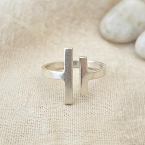 Puede incluir: Un anillo abierto de tono plateado con dos elementos de barra vertical de diferentes alturas que no se unen, creando un espacio en el centro de la banda. El metal tiene un acabado mate y liso con un diseño estructural y limpio.