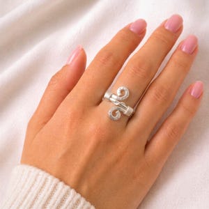 Può includere: Un anello in filo metallico color argento con un design a doppia spirale. La fascia è composta da più fili di metallo avvolti insieme, che formano due spirali distinte a estremità aperta che si sovrappongono leggermente al centro.