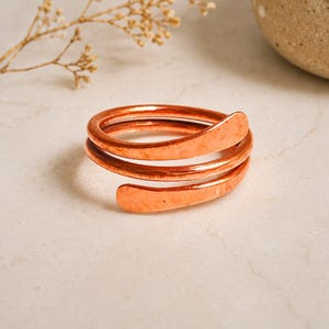 May include: A copper ring featuring a bypass design with three overlapping coils. The metal has a warm, metallic orange hue and a slightly hammered, textured finish on the flat, tapered ends of the band, creating a continuous spiral shape.