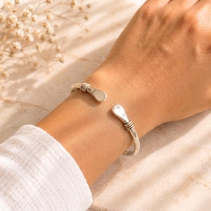 Puede incluir: Una pulsera tipo brazalete de tono plateado con un diseño abierto y dos extremos redondeados y aplanados. La banda presenta tres detalles de alambre fino enrollado horizontalmente cerca de cada extremo, con un acabado metálico.