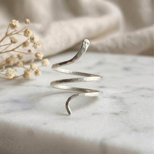 Può includere: Un anello in metallo color argento con un design a spirale simile a un serpente e una texture martellata. L'anello presenta un'estremità affusolata sulla parte superiore con due piccole rientranze che ricordano degli occhi, formando una spirale.