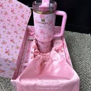 May include: A pink Stanley travel mug with a handle and a floral pattern, presented in a matching gift box. The box includes a pink satin bag. The mug has a clear lid and a gold-colored rim.