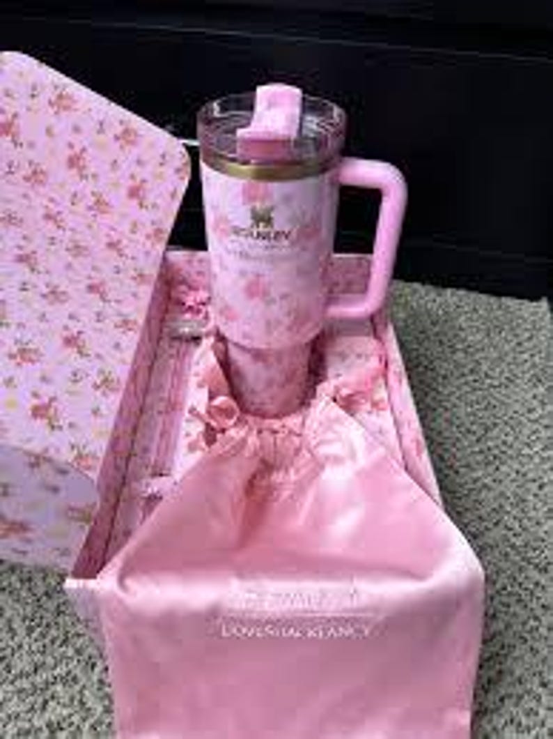 May include: A pink Stanley travel mug with a handle and a floral pattern, presented in a matching gift box. The box includes a pink satin bag. The mug has a clear lid and a gold-colored rim.
