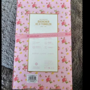 May include: Pink floral patterned box with the text "QUENCHER H2.0 TUMBLER". The box has a pink ribbon with the text "SERANINE | LOVE VACANCY". The background is a light pink with small pink flowers.