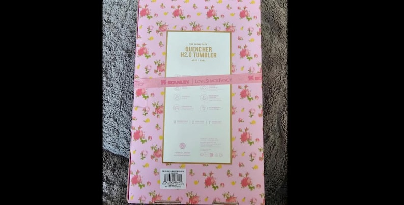 May include: Pink floral patterned box with the text "QUENCHER H2.0 TUMBLER". The box has a pink ribbon with the text "SERANINE | LOVE VACANCY". The background is a light pink with small pink flowers.