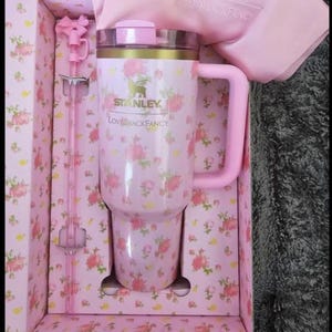 May include: A pink Stanley tumbler with a floral pattern, a pink handle, and a matching straw. The tumbler is in a floral-patterned box with the text "Stanley" and "Love Shack Fancy".