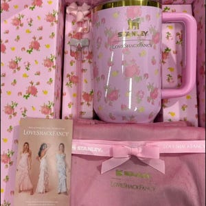May include: A pink floral Stanley LoveShackFancy mug with a handle, lid, and a matching pink gift box. The box contains a pink bow, a pink straw, and a pink pouch with a bow. A card with the text "Welcome to the World of LoveShackFancy" is also included.