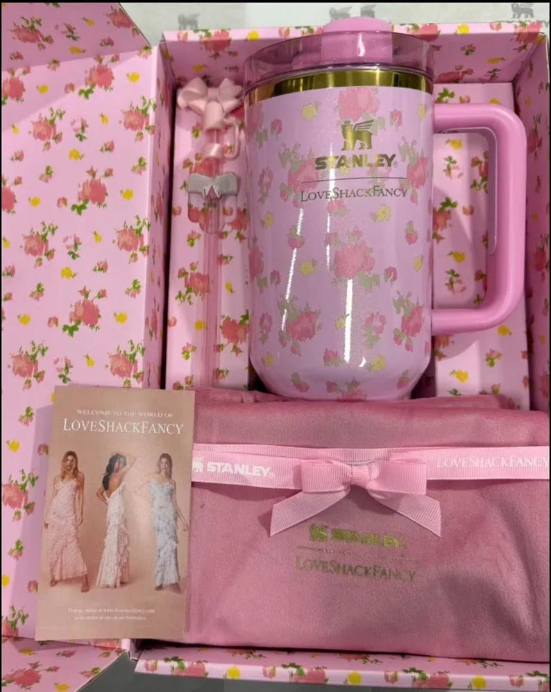 May include: A pink floral Stanley LoveShackFancy mug with a handle, lid, and a matching pink gift box. The box contains a pink bow, a pink straw, and a pink pouch with a bow. A card with the text "Welcome to the World of LoveShackFancy" is also included.