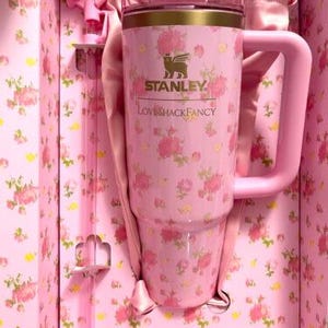 May include: A pink Stanley Loveshackfancy tumbler with a handle, floral pattern, and gold trim, presented in a matching floral-printed box. The tumbler has a capacity of approximately 40 ounces.