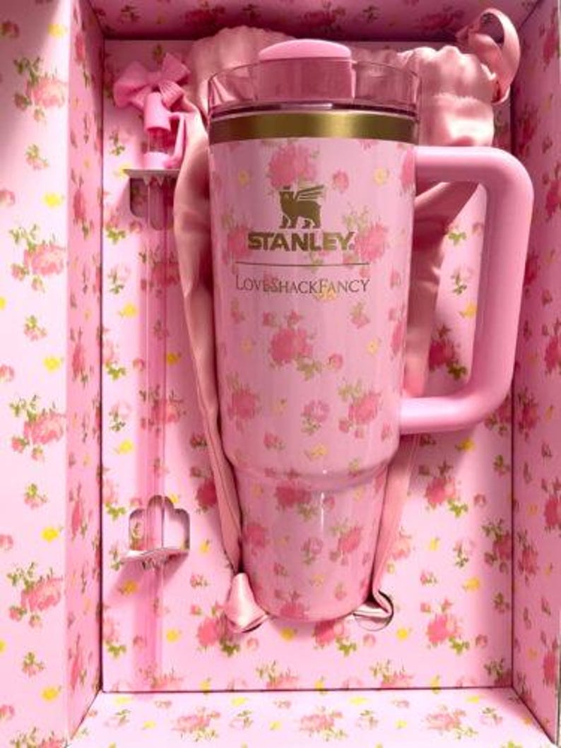 May include: A pink Stanley Loveshackfancy tumbler with a handle, floral pattern, and gold trim, presented in a matching floral-printed box. The tumbler has a capacity of approximately 40 ounces.