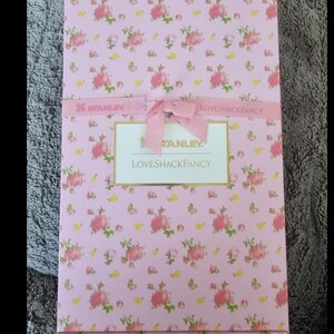 May include: A rectangular box with a pink floral pattern and a pink ribbon tied in a bow. The box has a label that reads "STANLEY LOVESHACKFANCY". The floral pattern includes pink roses and green leaves.