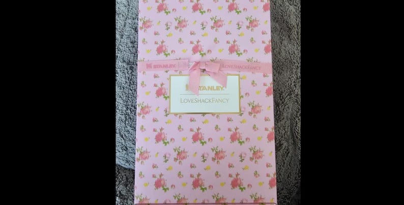 May include: A rectangular box with a pink floral pattern and a pink ribbon tied in a bow. The box has a label that reads "STANLEY LOVESHACKFANCY". The floral pattern includes pink roses and green leaves.