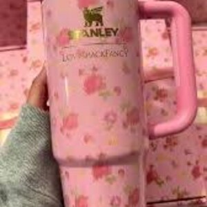 May include: Pink Stanley tumbler with a floral pattern and a handle. The tumbler features the "Stanley" logo and the text "LOVESHACKFANCY" in gold. The tumbler is held by a person wearing a gray sweater.