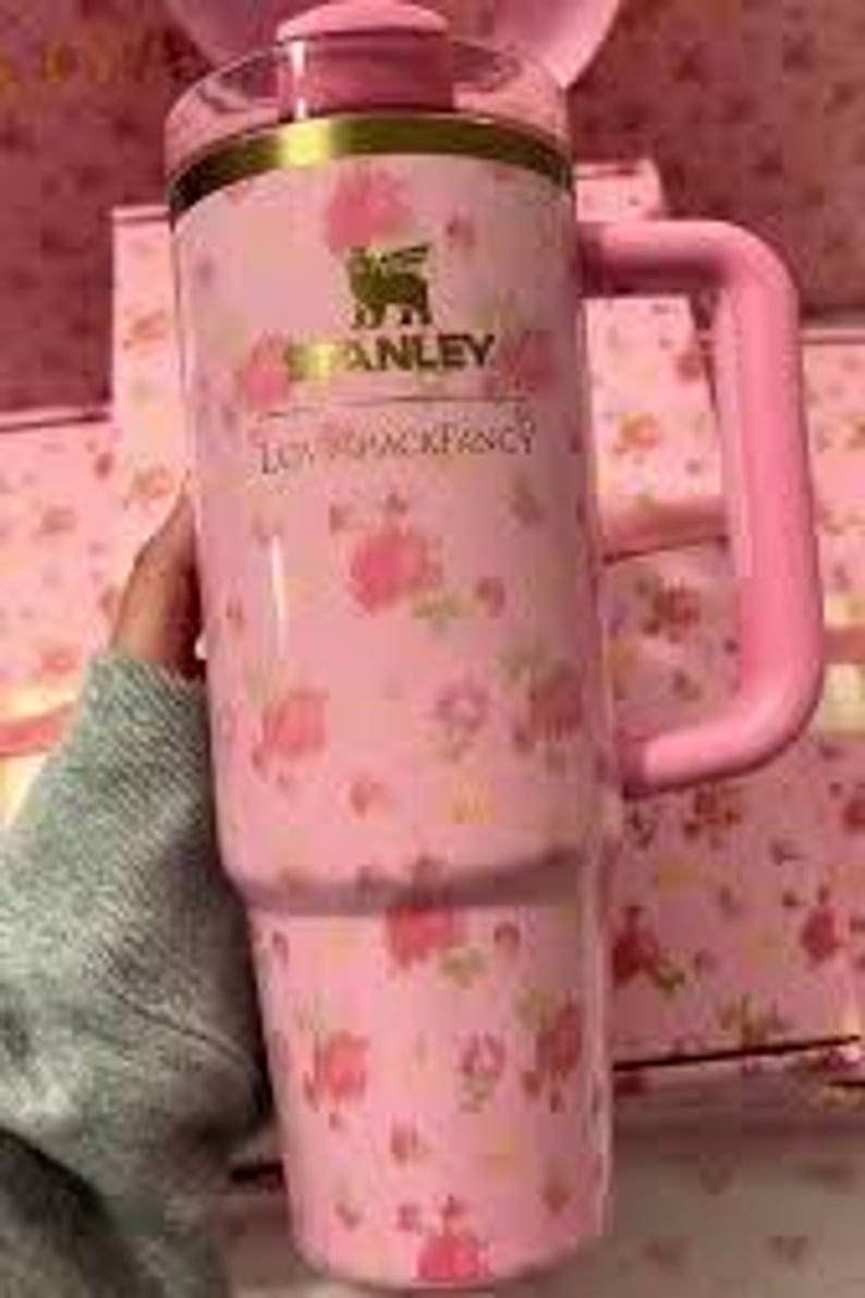 May include: Pink Stanley tumbler with a floral pattern and a handle. The tumbler features the "Stanley" logo and the text "LOVESHACKFANCY" in gold. The tumbler is held by a person wearing a gray sweater.