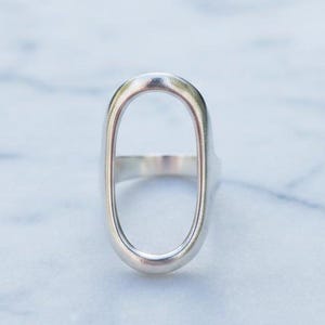 May include: A silver ring with an elongated oval design. The ring's band is smooth and connects to the oval shape, creating an open center. The ring is displayed on a marble surface.