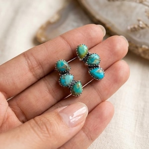 May include: A pair of turquoise and silver earrings. Each earring features three teardrop-shaped turquoise stones set in a silver frame. The earrings are displayed on a neutral background.