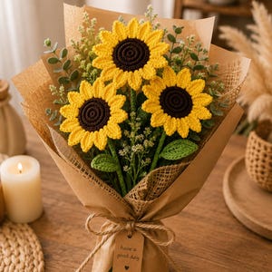 Puede incluir: Un ramo de tres girasoles de ganchillo con pétalos de color amarillo brillante y centros de color marrón oscuro, acentuados con hojas de ganchillo verdes y pequeñas ramitas de vegetación. Las flores están envueltas en papel marrón con un lazo de yute y una etiqueta que dice have a good day.