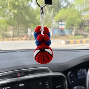 May include: A crocheted Spider-Man figure hanging from a silver metal keyring and a white cord. The figure features a red and blue textured yarn body with a round head, large white triangular eyes, and black stitched web detailing on the face.