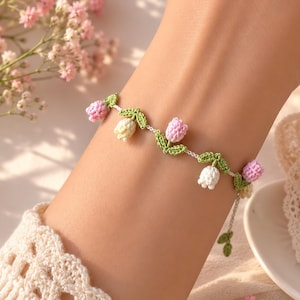 Puede incluir: Una pulsera delicada con pequeños dijes de flores tejidas a ganchillo en tonos rosa, blanco y amarillo, acentuados con hojas verdes tejidas. Los elementos florales están unidos a una fina cadena de hilo blanco que rodea la muñeca.