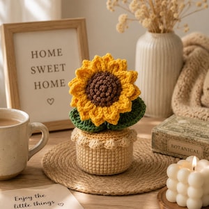 May include: A crocheted sunflower with bright yellow petals and a dark brown centre, potted in a beige textured container with a scalloped rim. The sunflower features two dark green leaves at the base, all crafted from yarn in a detailed stitch pattern.
