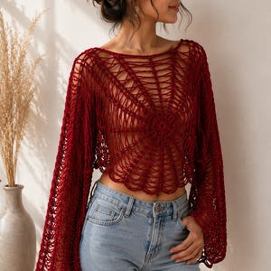May include: A deep red, long-sleeved crochet top featuring an intricate, open-knit spiderweb pattern that radiates from a central circular motif. The garment has a wide, boat-style neckline and loose, flared sleeves, paired with light blue denim jeans.