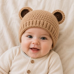 May include: A light brown crochet beanie featuring two rounded bear ears on top. The hat has a ribbed brim and a textured knit pattern. The ears are accented with a darker brown circular centre, creating a distinct bear-like appearance for the head.