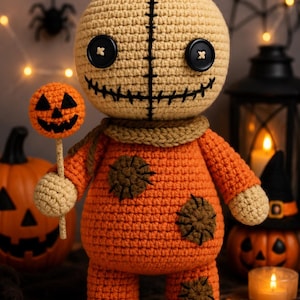 Puede incluir: Una muñeca de ganchillo con cabeza color canela, ojos de botón negros y boca cosida. Viste un mono naranja con parches circulares marrones y una bufanda color canela. La muñeca sostiene una pequeña piruleta de calabaza naranja.
