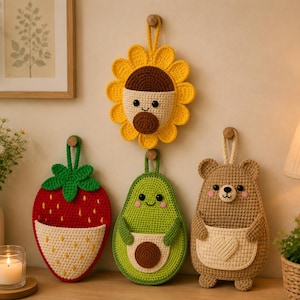 May include: Three crocheted wall hangings featuring a yellow sunflower, a red strawberry, and a brown bear, each with a small pocket on the front. The sunflower has a brown center, the strawberry has green leaves, and the bear has a cream heart pocket.