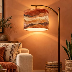 Peut inclure: Un lampadaire arqué avec un pied en métal sombre et un abat-jour cylindrique. L'abat-jour présente un motif de paysage abstrait en couches dans des tons terre cuite, orange brûlé, crème et brun foncé, diffusant une lueur douce et chaude.