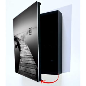 May include: Black and white framed artwork featuring a wooden pier extending into calm water. The artwork is mounted on a black frame, with a white background. The image depicts a serene landscape with a pier and a small structure.