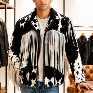 Könnte beinhalten: Schwarz-weiße Lederjacke mit Kuhfellmuster und langen weißen Fransen. Die Jacke hat einen klassischen Kragen und wird über einem grauen T-Shirt getragen. Weitere Lederjacken sind im Hintergrund zu sehen.