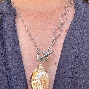 May include: A silver necklace with a natural seashell pendant. The shell is cream and brown with gold detailing. The necklace has a toggle clasp and a chain with oval links.