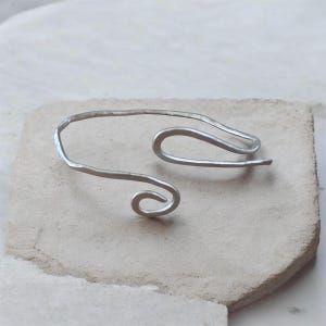Op de afbeelding: Een zilverkleurige metalen armband met een open ontwerp en een gehamerde textuur. De armband heeft een doorlopende, draadachtige vorm die aan één uiteinde in een spiraal krult en aan het andere uiteinde een afgeronde haakvorm heeft.