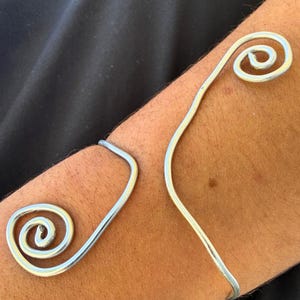 Op de afbeelding: Een zilveren armmanchet met een spiraalvormig ontwerp. De manchet wikkelt zich om de arm met twee spiraalaccenten. Het metaal glanst en reflecteert licht. Het ontwerp is eenvoudig en elegant.