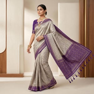 May include: A woman wearing a traditional saree. The saree is a light beige color with a subtle floral pattern and a deep purple border. The border features intricate designs and tassels. The blouse is a solid purple color, and the woman is wearing gold jewelry.