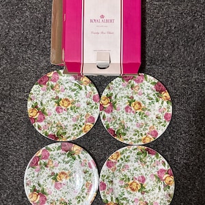 May include: Four round ceramic plates featuring a white base with a dense pattern of pink and yellow roses and green leaves. Above the plates is a rectangular cardboard box with a bright pink center panel labeled Royal Albert Country Rose.