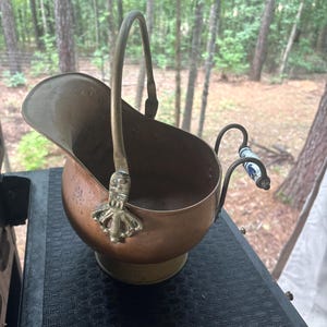 May include: A copper coal scuttle with a brass swing handle and a decorative brass mount at the base of the handle. The vessel features a secondary handle on the back with a white ceramic grip decorated with blue floral patterns and a brass base.