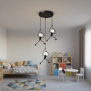 K&ouml;nnte beinhalten: Schwarze Metall-Pendelleuchte mit drei h&auml;ngenden Figuren, die jeweils eine kugelf&ouml;rmige Gl&uuml;hbirne halten. Die Figuren haben ein skurriles Design, das an Strichm&auml;nnchen erinnert. Die Leuchte ist an einer schwarzen Deckenplatte aufgeh&auml;ngt. Der Raum hat ein Kinderbett, einen Teppich und M&ouml;bel.